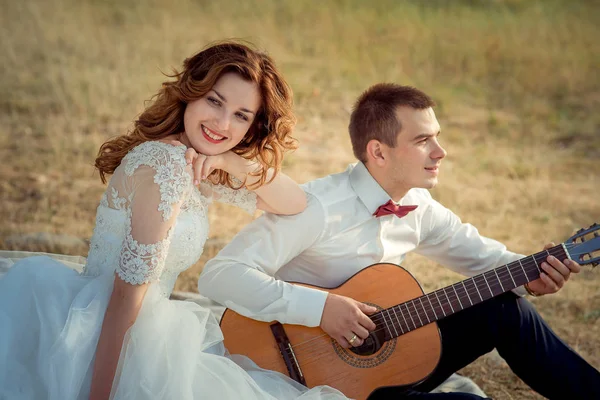 Portrait de la charmante mariée aux cheveux roux avec un joli sourire profitant du jeu de guitare de son marié élégant assis sur l'herbe . — Photo