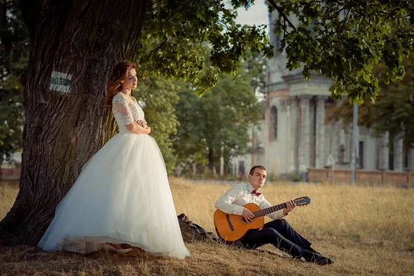 Matrimonio ritratto all'aperto. Il bel sposo sta suonando la chitarra mentre la sua affascinante sposa dai capelli rossi sta ascoltando e appoggiata all'albero . — Foto Stock