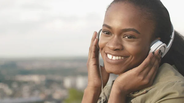 迷人的微笑和自然的美丽的非洲女孩的肖像在城市的模糊背景下通过她的耳机听音乐. — 图库照片