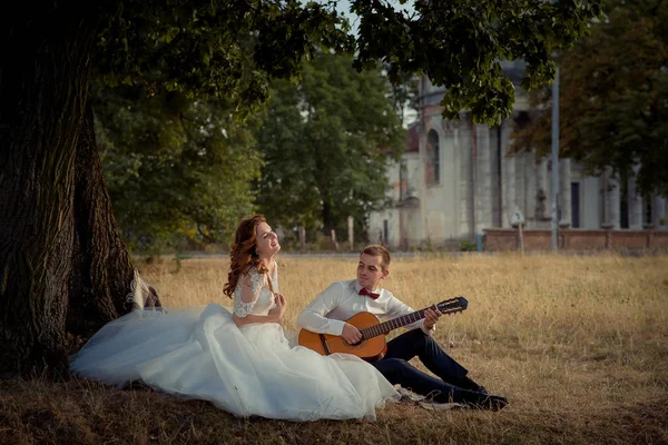 Ritratto degli sposi novelli e belli felici. Lo sposo sta suonando la chitarra e la sposa è seduta vicino sull'erba e gode . — Foto Stock