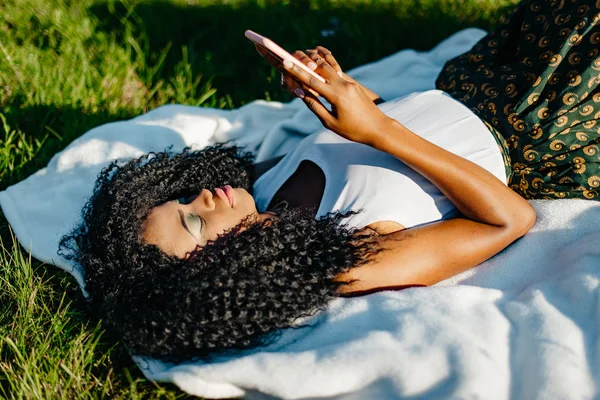 Hezká mladá Africká dívka je odpočívat na trávě. Ona je textilie a procházení prostřednictvím mobilního telefonu. Výše portrét. — Stock fotografie