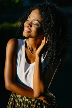 Güzel genç Afro-Amerikan kız çekici gülümseme ve yeşil göz gölgeler ile uzun siyah kıvırcık saçları çok dokunaklı ve güneşli bahçedeki Kapalı gözlerle zevk.