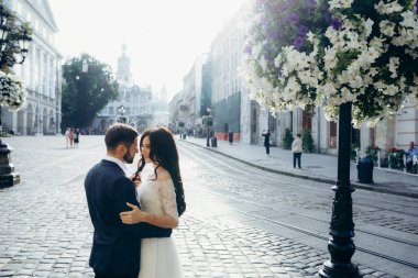 Hassas düğün portre güneşli sokakta. Yakışıklı damat usulca çiçeklerle süslenmiş sokak lambası yakınındaki esmer gelin saç dokunuyor.