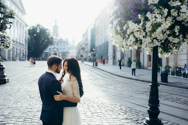 Sensitive wedding portrait in the sunny street. Handsome groom is softly touching the hair of his brunette bride near the street lamp decorated with flowers. — Stock Photo, Image