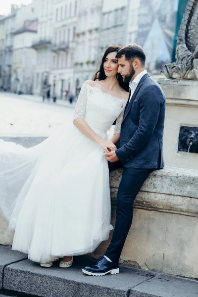 Das charmante Brautpaar umarmt sich sanft, während es auf dem alten Brunnen sitzt. Hochzeitsfoto in voller Länge. — Stockfoto