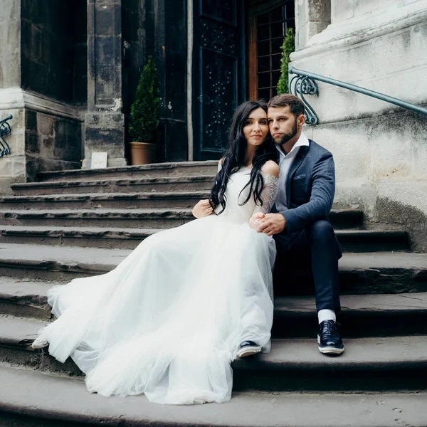 La elegante pareja recién casada feliz está tiernamente abrazando y tomándose de la mano mientras se sienta en las escaleras de la antigua iglesia barroca . — Foto de Stock