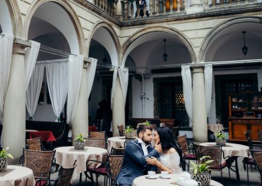 Güzel yeni evli çiftin açık restaursnt masada oturuyor. Yakışıklı damat usulca esmer gelinin yüzünü okşayarak.