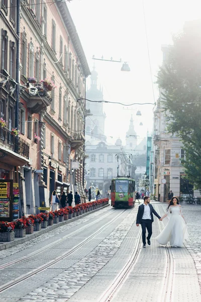 Full-length colpo di nozze della coppia di sposi alla moda che si tiene per mano mentre si cammina lungo la strada della città . — Foto Stock