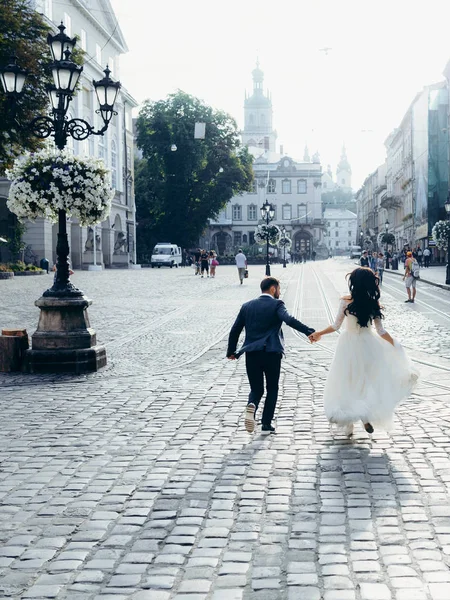 Das fröhlich-schöne Brautpaar hält Händchen, während es die sonnige Stadtstraße entlangläuft. vertikale Rückansicht. — Stockfoto
