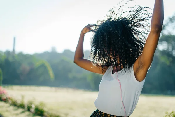 Активная молодая хорошенькая африканская девушка эмоционально танцует и трясет своими темными вьющимися волосами, слушая музыку в наушниках на солнечном лугу . — стоковое фото