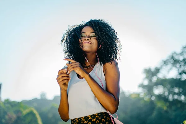 Outdoor Portret van de jonge mooie Afrikaanse meisje met lang donker krullend haar genieten van muziek in haar oortelefoons met gesloten ogen, charmante glimlach en natuurlijke make-up. — Stockfoto