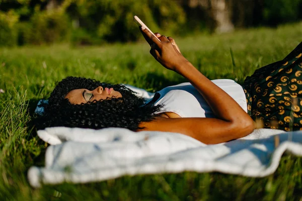 Krásná Africká dívka s zelené oční stíny je položil na trávník a chatování, posílání SMS zpráv a prohlížení prostřednictvím mobilního telefonu. Boční pohled. — Stock fotografie