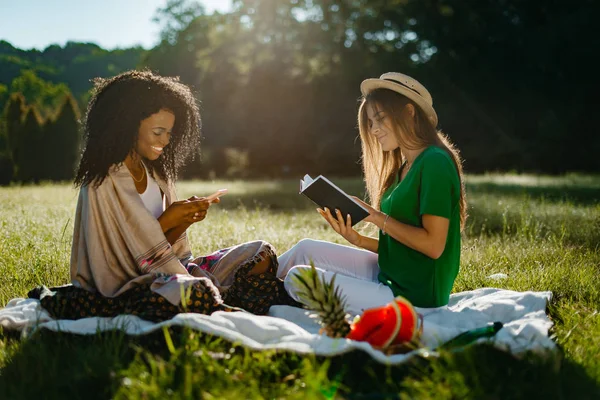 Piknik dvou více závod dívka přátel. Krásná mladá blondýnka je čtení knihy, zatímco její šťastný usmívající se africký přítel je chatování a procházení přes mobilní telefon. — Stock fotografie