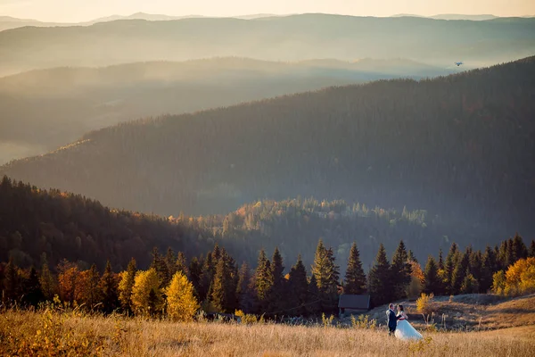 Magnifica vista sul paesaggio delle montagne nebbiose gokden durante il tramonto. Allegro glamour coppia di sposi sta ballando sul prato . — Foto Stock