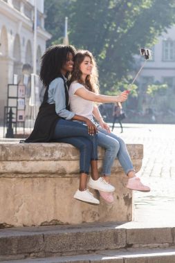 Çeşme ve selfie stick kullanarak alma selfies oturan güzel kız tam uzunlukta görünümü.