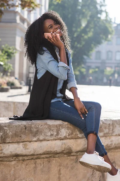 Visão completa da menina afro-americana elegantemente vestido falando via telefone celular e rindo. Ela está sentada na fonte no centro da cidade . — Fotografia de Stock