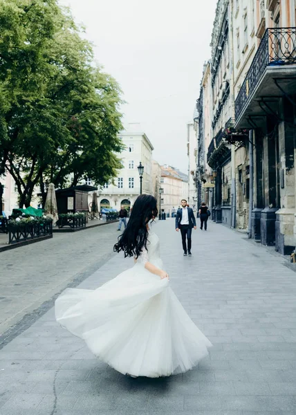 Full-length portrait of the charming brunette bride spinning round in her long wedding dress in the sunny street. — Stock Photo, Image