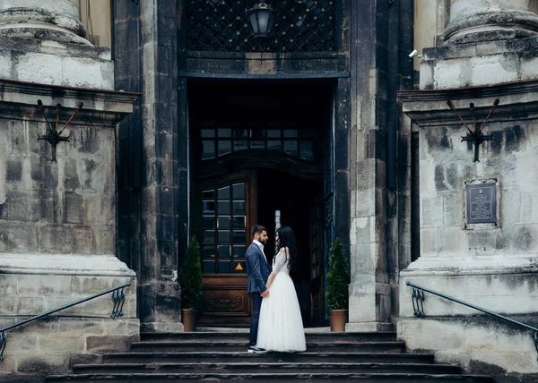 Yeni evli genç çift sevimli yumuşak el ele tutuşarak ve birbirine Barok kilisenin eski merdivenlerde dururken bakarak.