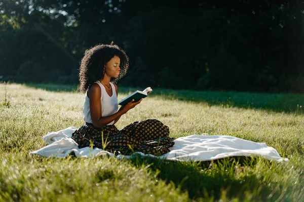 Během pikniku na slunné louce je relaxační s knihou okouzlující Africká dívka s přírodní make-up a kudrnaté vlasy. — Stock fotografie