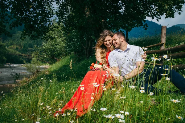 Feliz casal rindo é ternamente abraçando enquanto sentado no prado de camomila sob a árvore perto do rio e cerca de madeira. A mulher loira bonita com o buquê . — Fotografia de Stock