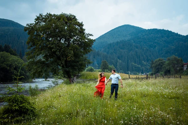 Portrait of the beautiful happy couple holding hands and walking along daisy meadow near the river in the mountains covered with green trees.