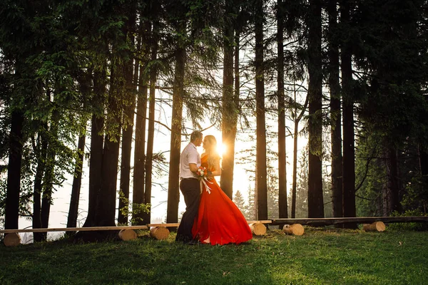 夕日の中に森で優しく抱いてエレガントなカップルのフルレングスのビュー。長い赤 drrress の女の子は、野生の花の花束を保持しています。. — ストック写真