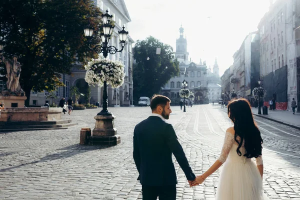 Vista trasera de media longitud de la hermosa pareja de recién casados felices tomados de la mano en la soleada calle de la ciudad . — Foto de Stock