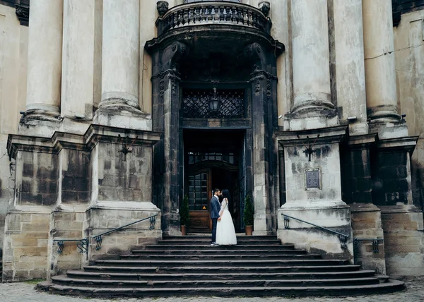 Horizontal full-length wedding portrait of the beautiful attractive newlywed couple holding hands and rubbing noses at the stairs of the old baroque church. — Stock Photo, Image