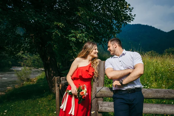 Close-up retrato emocional do atraente jovem casal feliz falando enquanto se apoia na cerca de madeira no prado margarida perto do rio nas montanhas . — Fotografia de Stock