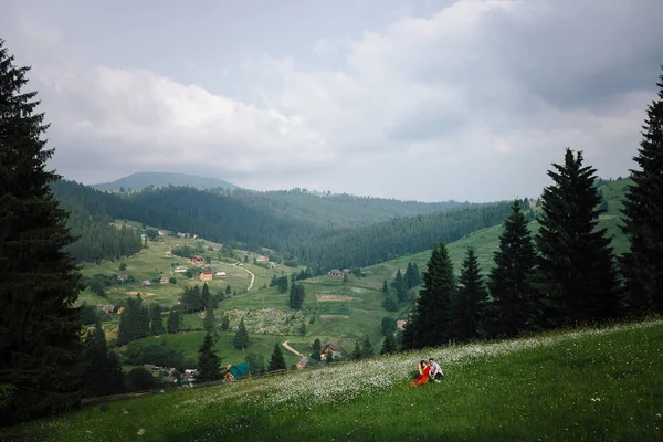 Panorama krásný výhled hory, krajina a louce pokryté sedmikrásky. Krásný pár sedí na rozkvetlé louce heřmánek. — Stock fotografie