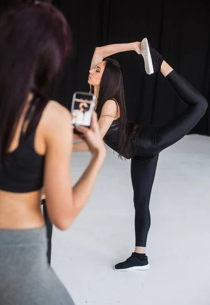 Pandangan belakang kabur dari wanita atletik mengambil foto teman gadis olahraganya melakukan busur berdiri di latar belakang hitam . — Stok Foto