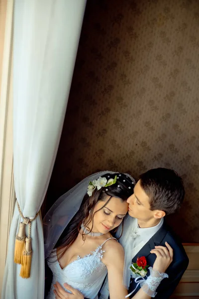 Über romantischem Hochzeitsporträt. der hübsche Bräutigam umarmt und küsst seine schöne brünette Braut zärtlich auf die Stirn im alten, reichen Zimmer. — Stockfoto
