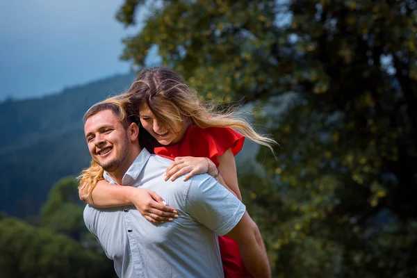 The handsome happy man is giving the piggyback ride his charming smiling blonde girlfriend at the background of the sunny green mountains. — Stock Photo, Image