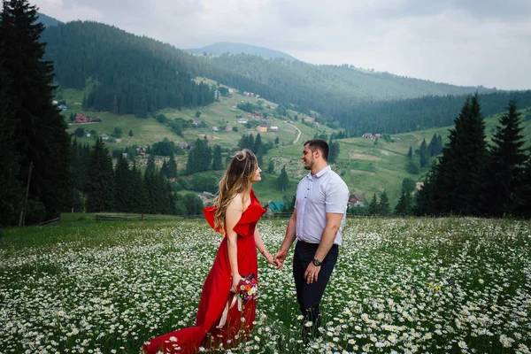 Outdoor portrait of the attractive smiling couple holding hands on the daisy meadow at the background of the beautiful green mountains. The girl is holding the bouquet of wild flowers.