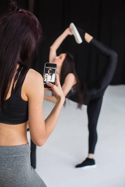 The back view of the sports woman taking photo of the blurred athletic girl in the pose of standing bow. The view of the girl friend on the phone. — Stock Photo, Image