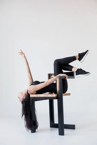 The sports attractive flexible woman in sportwear is stretching on the chair with the legs on the chair back at the white background. — Stock Photo, Image