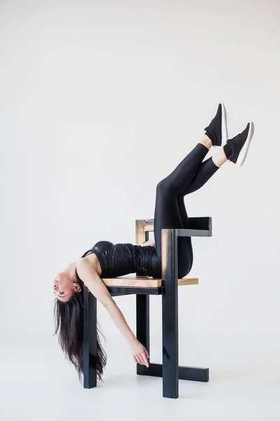 Full-length side portrait of the athletic young woman in sportsuit stretching with the legs on the chair back at the white background. — Stock Photo, Image