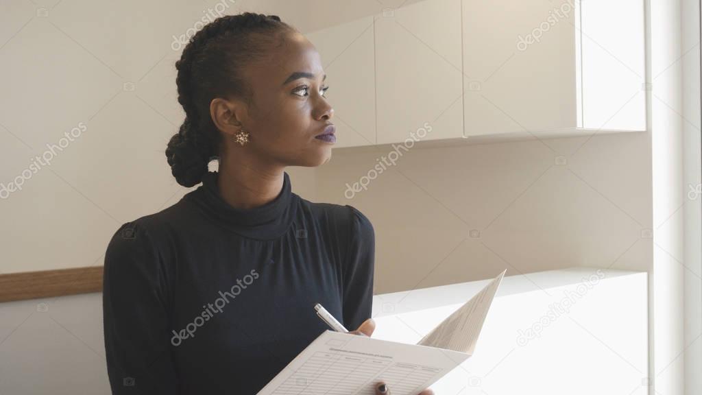 Charming serious african woman with natural make-up is thoughtfully looking aside while holding the documents in the office. Girl at the reception.