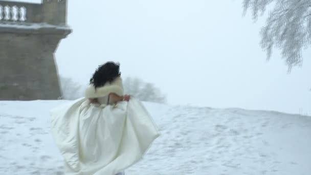 The back view of the young bride with long dark curly hair in long wedding dress running along the snowy meadow. No face. — Stock Video