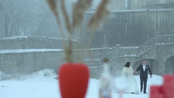 The stylish newlyweds are softly holding hands while walking along the snowy meadow at background of old house. Focus on the table decorated with wine glasses, candles, herbs and vase with wheats. — Stock Video