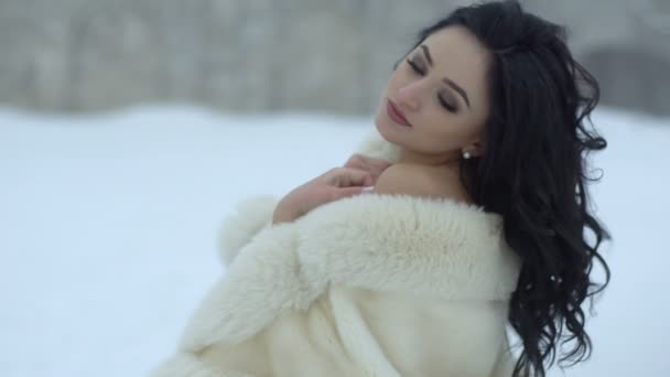 Das Nahaufnahme-Porträt der zarten Braut mit langen dunkelgelockten Haaren im Pelzmantel, die während des Schneefalls auf der Wiese in die Kamera blickt. — Stockvideo