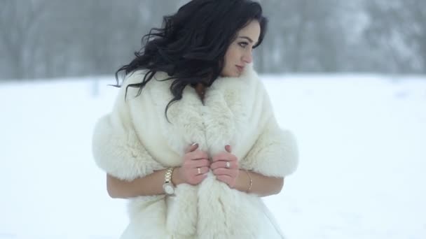 Portrait of the young charming bride with beautiful long dark curly hair wrapping in fur coat, looking away and then in camera. Snowy meadow location — Stock Video