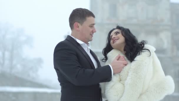 Romantic wedding portrait of the happy young couple of newlyweds holding hands and tenderly looking in each other eyes. The handsome groom is gently kissing his lover in the nose during the snowfall. — Stock Video