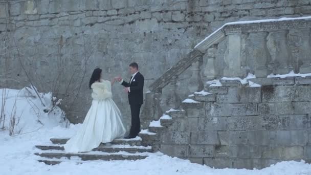 The attractive brunette bride is going up the stairs with the held of groom hand. They are tenderly kissing on the snowy stairs of the old castle. — Stock Video