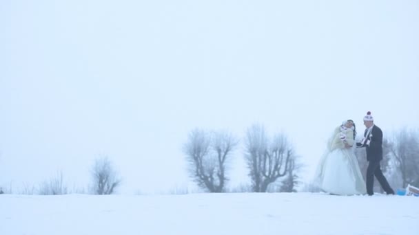 Full-length shot of the happy adorable newlyweds in wedding clothes and funny hats happily talking, laughing and walking with the cups and drinking tea along the snowy meadow. — Stock Video