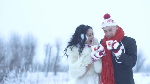 Glada skratt bedårande nygifta i bröllop kläder, roliga handskar och hattar dricka te eller kaffe via koppar på snöiga ängen. — Stockvideo