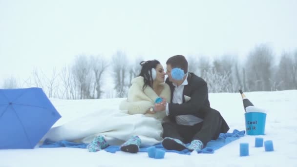 Winter blue picnic for beautiful newlywed couple. They are sitting on the plaid near the umbrella, candles and champagne in bucket while tenderly hugging, holding hands and rubbing noses. — Stock Video