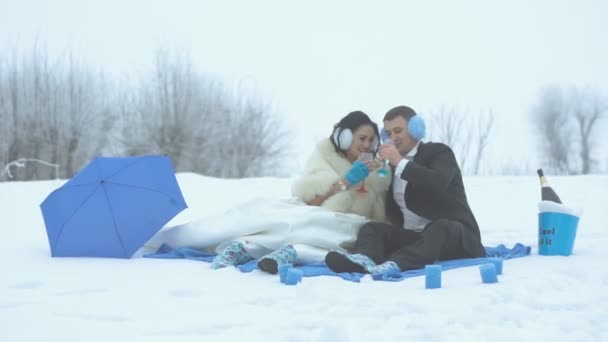 Heureux jeunes mariés rient en buvant du champagne lors de leur pique-nique d'hiver de mariage de couleur bleue décoré avec parapluie et bougies . — Video
