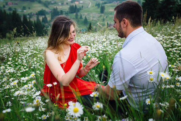 Menina loira bonita com sorriso bonito e maquiagem natural está jogando o jogo que ele me ama, me ama não na margarida enquanto sentado na frente de seu namorado no prado de camomila florescendo . — Fotografia de Stock