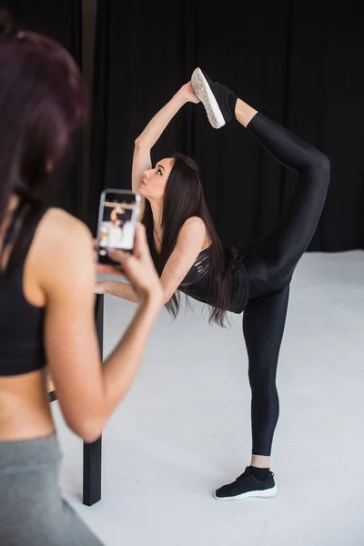 Full-length view of the athletic young sportwoman in the standing bow pose while her friend is taking photo of her on the mobile phone in the studio. — Stock Photo, Image
